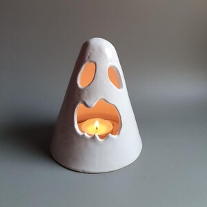 May include: White ceramic ghost candle holder with a cut-out face and a lit tea light inside.