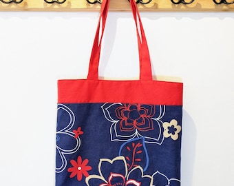 May include: A navy blue tote bag with a red top and handles, adorned with a floral pattern in white, red, and blue. The bag is hanging on a wooden rack with black hooks, against a white wall.