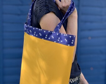May include: A yellow tote bag with a blue and white patterned trim. The bag has a wide, flat base and two handles.