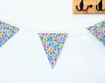 May include: A bunting banner featuring three triangular fabric flags attached to a white ribbon. The fabric has a light blue background with a repeating pattern of small yellow, pink, and teal flowers with dark centers and green leaves.