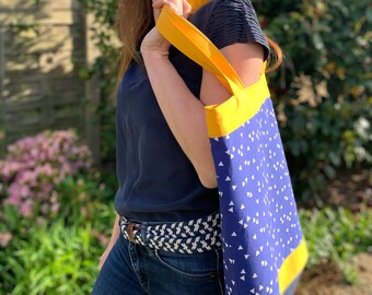 May include: A woman is carrying a blue and yellow tote bag with a white triangle pattern. The bag has yellow straps and a yellow stripe across the bottom.