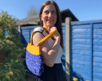 May include: A woman wearing a white and black top and a black skirt is holding a blue and yellow tote bag with white triangles. The bag has a yellow strap.