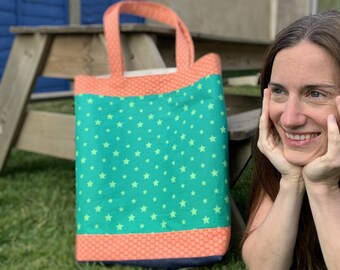 May include: A green tote bag with white stars and an orange and navy blue trim. The bag is sitting on a wooden bench with grass in the background.