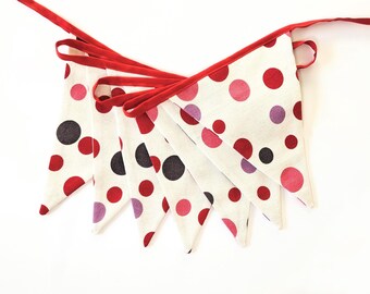 May include: A fabric bunting with white pennants adorned with red, pink, purple, and black polka dots. The edges are trimmed with red fabric, and a red ribbon runs along the top for hanging. Great for parties and celebrations.