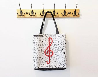 May include: White tote bag with a musical note pattern and a red treble clef design. The bag has black handles and a black bottom trim. The bag is hanging on a wooden rack with black hooks.