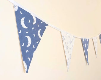 May include: A blue and white fabric bunting with a moon and star pattern. The bunting is strung up on a white string and is hanging on a white wall.