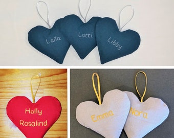 May include: Assortment of personalized heart-shaped fabric ornaments in various colors, including red, blue, and gray. Each heart features a name embroidered in gold thread. The names include "Holly Rosalind", "Lotti", "Libby", "Laila", "Emma", and "Nora".