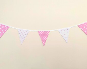 May include: A pink and white fabric banner with triangular flags. The flags are strung on a white ribbon. Some flags have a pink background with white patterns, while others have a white background with pink patterns.