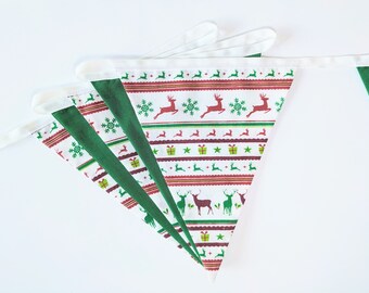 May include: Christmas-themed bunting with a festive pattern. The fabric features reindeer, snowflakes, and gift boxes in red, green, and white. The bunting is made of triangular flags with a green border and white trim.