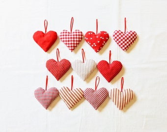 May include: Twelve red fabric hearts with different patterns, including gingham, polka dots, and stripes. The hearts are hanging from a white background.