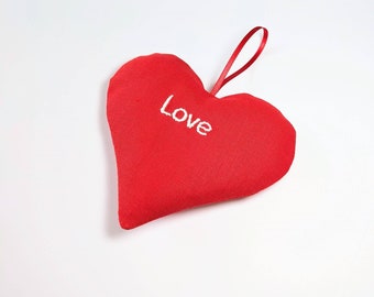 May include: A red heart-shaped fabric ornament with the word "Love" embroidered in white. It has a red ribbon loop for hanging. The ornament is a Valentine's Day or romantic-themed decoration.