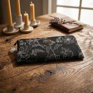 Könnte beinhalten: Schwarze Geldbörse mit weißem Blumenmuster, Reißverschluss und kleinem Lederetikett. Sie liegt auf einem Holztisch, im Hintergrund Kerzen und ein Buch. Das Muster zeigt Blumen und Blätter.