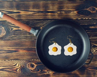 May include: A black cast iron skillet with two fried egg earrings inside. The earrings are gold with a white egg and a yellow yolk.