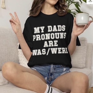 May include: A black short-sleeved t-shirt featuring white block lettering that reads MY DADS PRONOUNS ARE WAS/WERE. The shirt has a crew neckline and a standard fit, with the text centered prominently across the chest area in a bold, collegiate style font.