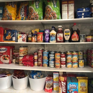 May include: A well-stocked pantry with shelves filled with various food items, including cereal boxes, bags of chips, canned goods, jars of sauces, and boxes of pasta. There are also white plastic bins on the bottom shelf.
