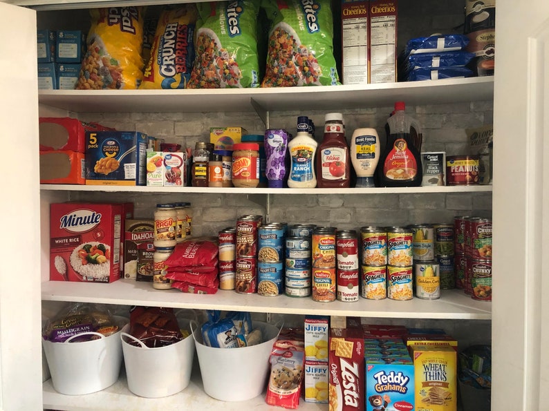 May include: A well-stocked pantry with shelves filled with various food items, including cereal boxes, bags of chips, canned goods, jars of sauces, and boxes of pasta. There are also white plastic bins on the bottom shelf.