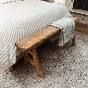 May include: A rustic wooden bench with a gray throw blanket on top, sitting on a gray and white patterned rug in front of a bed with white bedding.