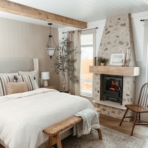 May include: A bedroom with a white bed, a wooden bench, a stone fireplace with a black electric fireplace insert, and a rug with a patterned design.