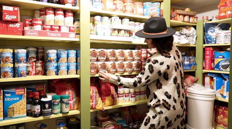 May include: A well-stocked pantry with shelves full of canned goods, pasta, and other food items. A person wearing a leopard print sweater and a black hat is reaching for a can of food on a shelf.