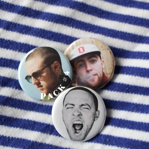 Puede incluir: Tres pines redondos con botones que muestran retratos de un hombre con una gorra de b&eacute;isbol, un hombre con barba y un hombre con la boca abierta. Los pines est&aacute;n sobre un fondo de tela a rayas azules y blancas. El texto "PACK #" es visible en uno de los pines.