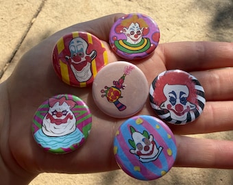Puede incluir: Cinco chapas redondas con alfiler que presentan ilustraciones de payasos coloridos y una chapa central con el gráfico de una pistola de juguete colorida. Los diseños incluyen varios rostros de payasos con patrones rojos, azules, verdes y amarillos.