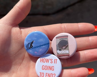 Puede incluir: Tres botones redondos. Uno es azul con la silueta de una persona subiendo una escalera. Otro es rosa con el texto "Mococoa" y un gráfico de cacao. El tercero es blanco con el texto "HOW'S IT GOING TO END?"