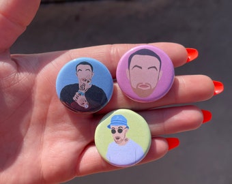 Puede incluir: Tres botones redondos con diferentes ilustraciones de un hombre con barba y pelo corto castaño. Los botones están sobre un fondo azul claro, morado claro y verde claro.