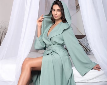 May include: A light green hooded robe with a long, flowing design. The robe is tied at the waist with a matching sash. The model is sitting on a bed with white linens and a patterned pillow.