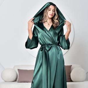 Könnte beinhalten: Ein langer, smaragdgrüner Satin-Bademantel mit Kapuze und Wickeldesign. Der Bademantel hat weite Ärmel und einen passenden Bindegürtel. Das Model trägt den Bademantel in einem Studio mit weißem Sofa und großem kreisförmigen Spiegel.