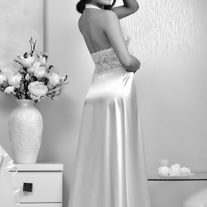 K&ouml;nnte beinhalten: Eine Frau tr&auml;gt ein wei&szlig;es Satin-Schlafkleid mit Spitzendetails. Das Kleid hat einen Halterneck und einen flie&szlig;enden Rock. Die Frau steht in einem Raum mit wei&szlig;en W&auml;nden und einem wei&szlig;en Nachttisch.
