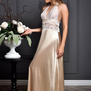 K&ouml;nnte beinhalten: Eine Frau tr&auml;gt ein wei&szlig;es Spitzenhaltertop und ein langes, flie&szlig;endes, champagnerfarbenes Satin-Schlafkleid. Sie steht vor einer grauen Wand mit wei&szlig;en Verzierungen und einem Blumenarrangement auf einem Tisch.