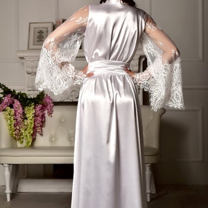 May include: A white satin robe with long sleeves featuring delicate lace detailing. The robe has a sash tie at the waist and a flowing skirt.