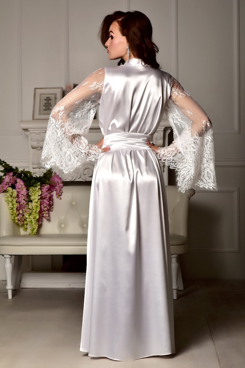 May include: A white satin robe with long sleeves featuring delicate lace detailing. The robe has a sash tie at the waist and a flowing skirt.