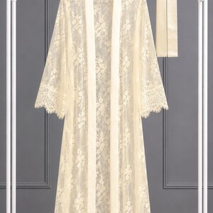 May include: A floor-length, cream-colored lace robe featuring a floral pattern, long sleeves with scalloped edges, and a solid satin-finish trim along the front opening. A matching long, narrow satin-finish sash is included with the garment.