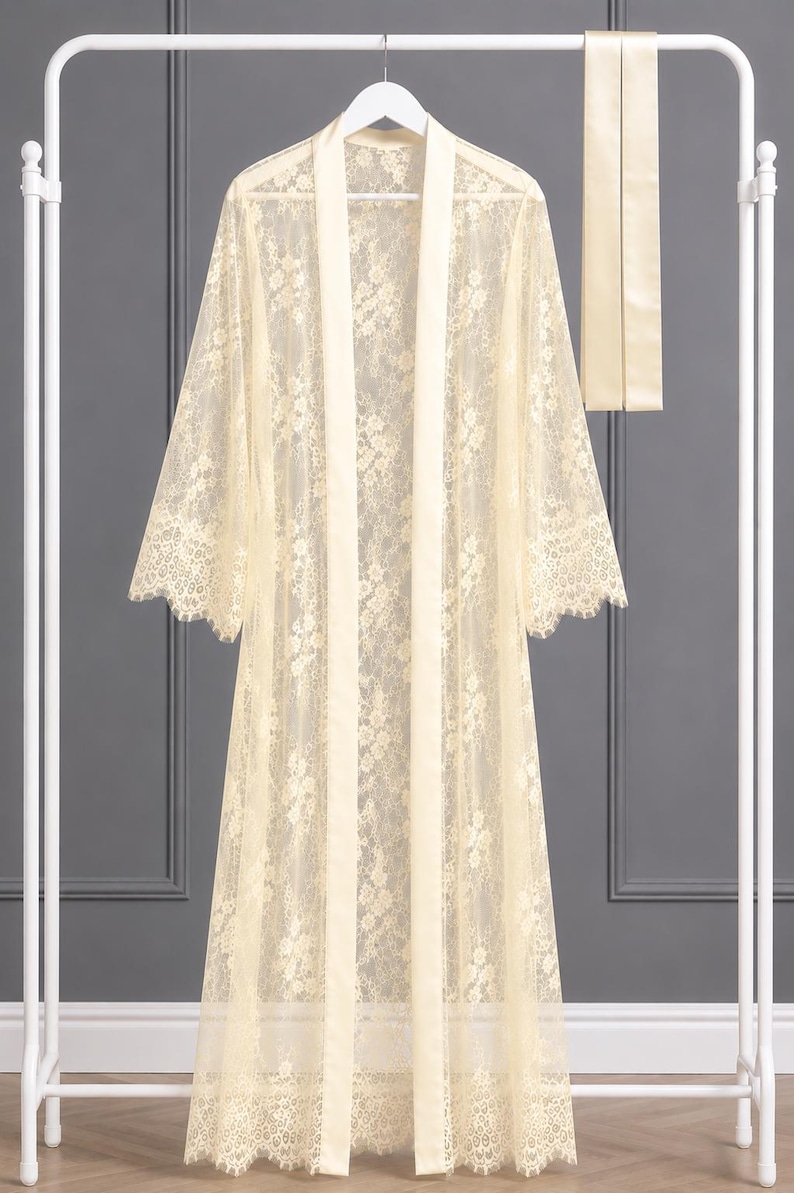 May include: A floor-length, cream-colored lace robe featuring a floral pattern, long sleeves with scalloped edges, and a solid satin-finish trim along the front opening. A matching long, narrow satin-finish sash is included with the garment.