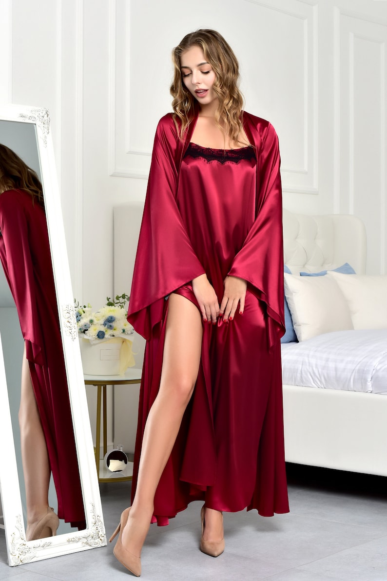 May include: A long, burgundy satin robe with a black lace trim. The robe has long sleeves and a deep V-neckline. The model is wearing the robe and standing in front of a mirror.