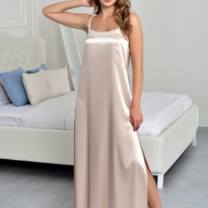 May include: A long, light beige satin slip dress with a lace trim at the neckline. The dress has a side slit and is worn by a model with long, wavy hair.