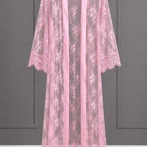 May include: A floor-length light pink robe crafted from sheer floral lace. The garment features long, wide sleeves with scalloped edges, a smooth satin-finish trim along the front opening, and a matching scalloped hemline at the bottom.