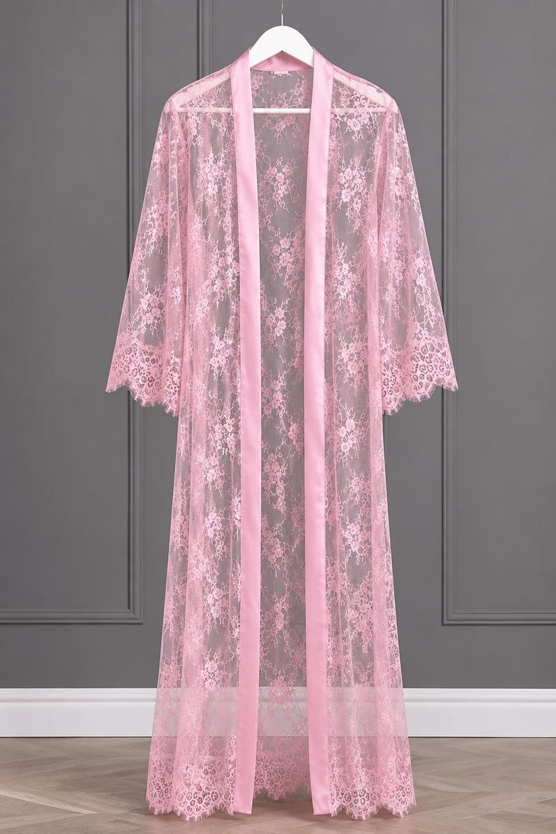 May include: A floor-length light pink robe crafted from sheer floral lace. The garment features long, wide sleeves with scalloped edges, a smooth satin-finish trim along the front opening, and a matching scalloped hemline at the bottom.
