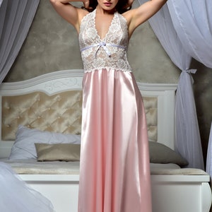 May include: A woman wearing a white lace and pink satin nightgown. The gown has a halter neckline and a bow at the bust. She is standing in front of a white bed with a white headboard.