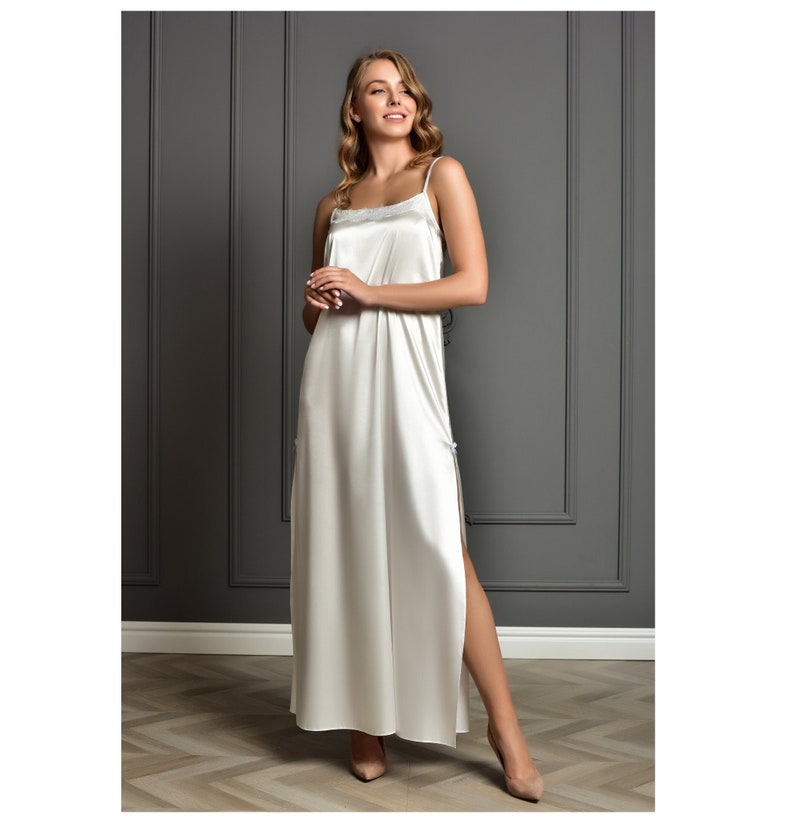 May include: A white satin slip dress with thin straps and a lace trim at the neckline. The dress has a side slit and falls to the ankles.