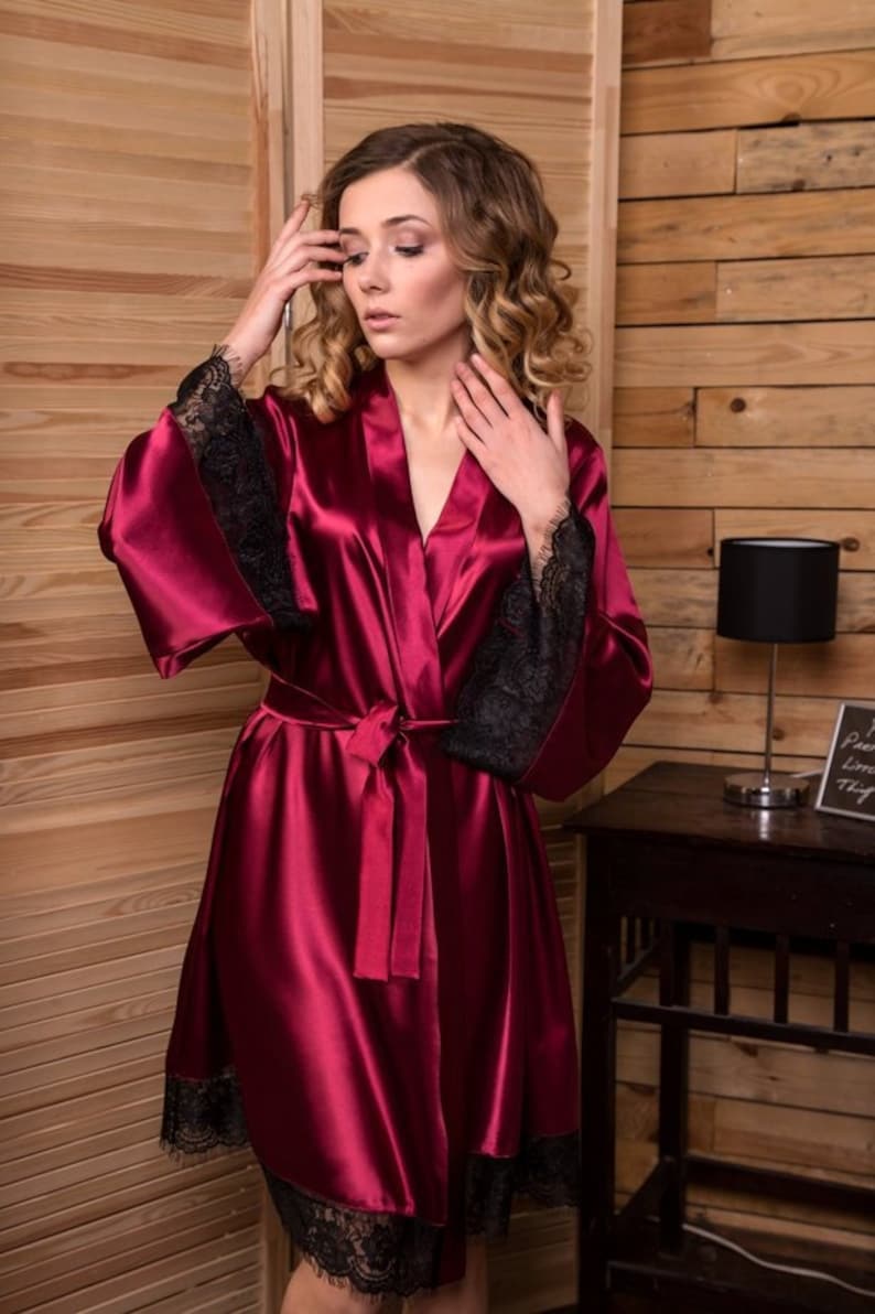 May include: A burgundy satin robe with black lace trim on the sleeves and hem. The robe has a tie belt and a V-neck.