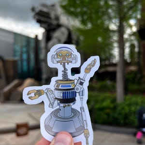 May include: A white, blue, and gold robot sticker with a spring body and a round base. The robot has two arms, one with a claw and the other with a long, thin arm. The robot is holding a sign that says "Welcome to Batuu".