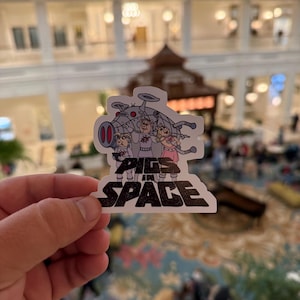 May include: A clear sticker featuring the text "Pigs in Space" with a cartoon image of three pigs in a spaceship. The pigs are wearing space suits and are in front of a gray spaceship with red accents.