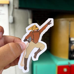 May include: A sticker of a cartoon character wearing a yellow helmet and brown suit, flying with a jetpack. The character is flying with their arm outstretched and their legs bent.