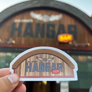 May include: A sticker of a brown wooden building with a red roof and the text "Hangar Bar" in white letters. The sticker is held in a hand in front of a larger version of the same building.