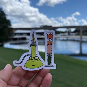 Könnte beinhalten: Zwei Cartoon-Aufkleber: ein gelber Bunsenbrenner mit blauer Flamme und ein Becherglas mit Gesicht. Die Aufkleber werden vor einem malerischen Hintergrund aus Wasser, Bäumen und einer Brücke unter einem bewölkten Himmel gehalten.