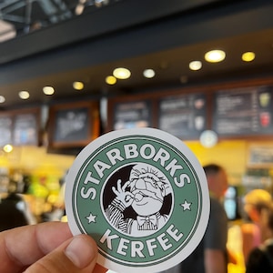 Könnte beinhalten: Ein runder Aufkleber mit grünem Rand und den weißen Worten "STARBORKS KERFEE". Der Aufkleber zeigt eine Zeichentrickfigur mit weißer Kochmütze und Fliege, die eine "okay" Handgeste macht.