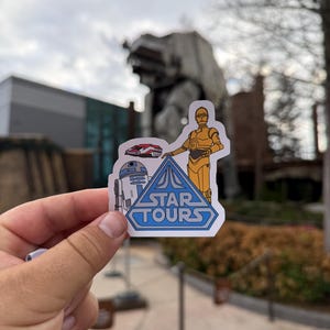 Könnte beinhalten: Ein Star Tours-Aufkleber mit C-3PO, R2-D2 und einem roten Fahrzeug. Der Aufkleber hat ein blaues Dreieck mit den Worten "STAR TOURS" und wird vor einem großen AT-AT-Walker und einem bewölkten Himmel gehalten.