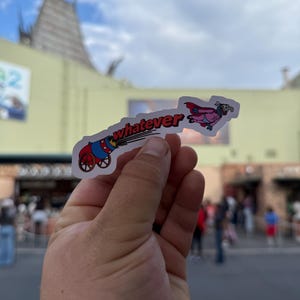 Op de afbeelding: Een kleurrijke sticker met een cartoonkanon met het woord "whatever" en een vliegend personage. De sticker wordt omhoog gehouden tegen een vage buitenachtergrond, met een bewolkte lucht en gebouwen zichtbaar.